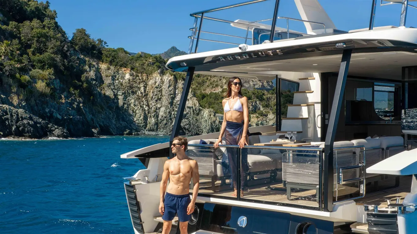 A man and woman stand on the deck of a luxury yacht named 'Absolute', with clear blue water and mountainous scenery in the background.