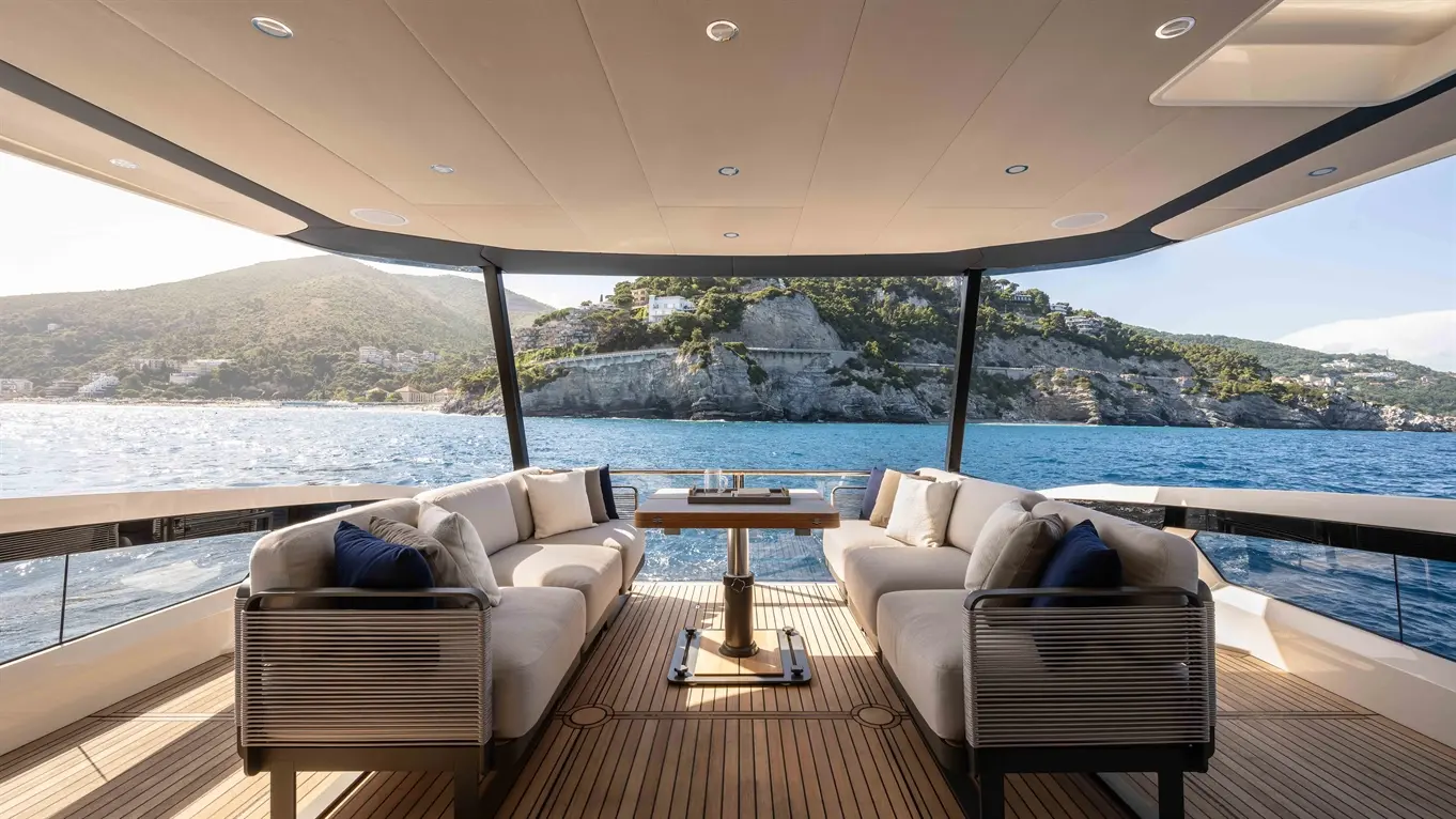 Luxurious yacht deck with comfortable seating and a table, overlooking clear blue waters and a scenic coastal backdrop.