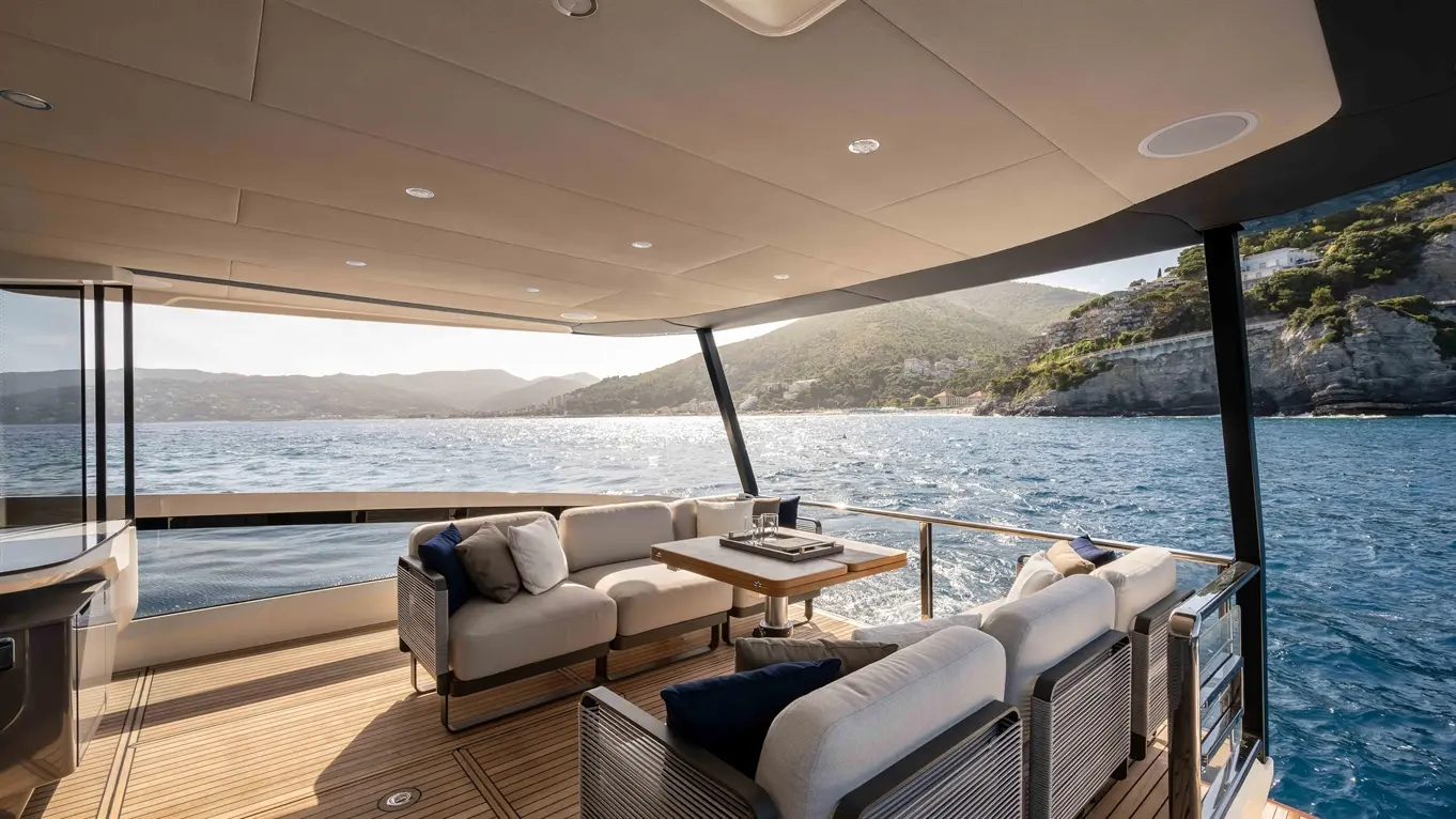 Luxurious yacht deck with stylish seating and a table, overlooking sparkling water and mountainous coastline in bright sunlight.