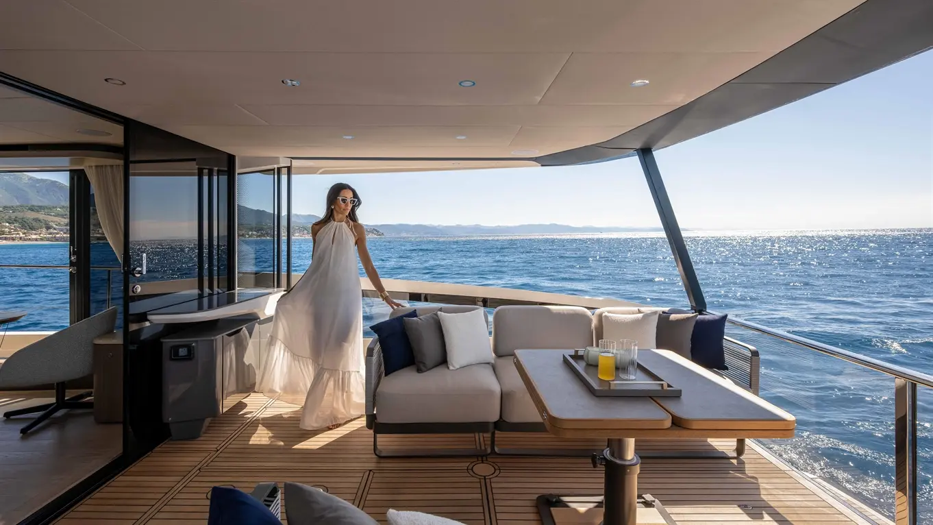 A woman in a flowing white dress stands on a luxury yacht's deck, overlooking a sparkling sea. Sunlight glistens.