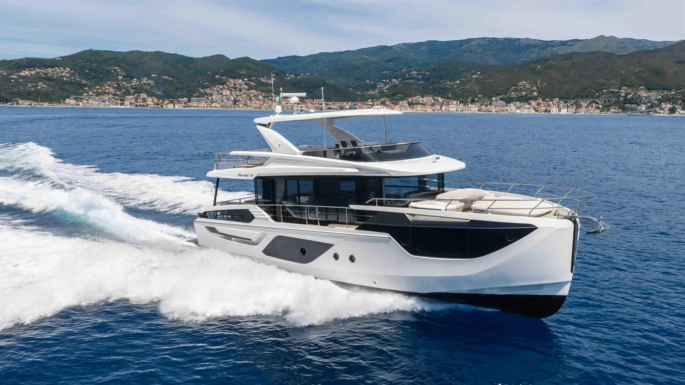 A modern yacht navigates through clear blue waters with a coastal town and green hills in the background.