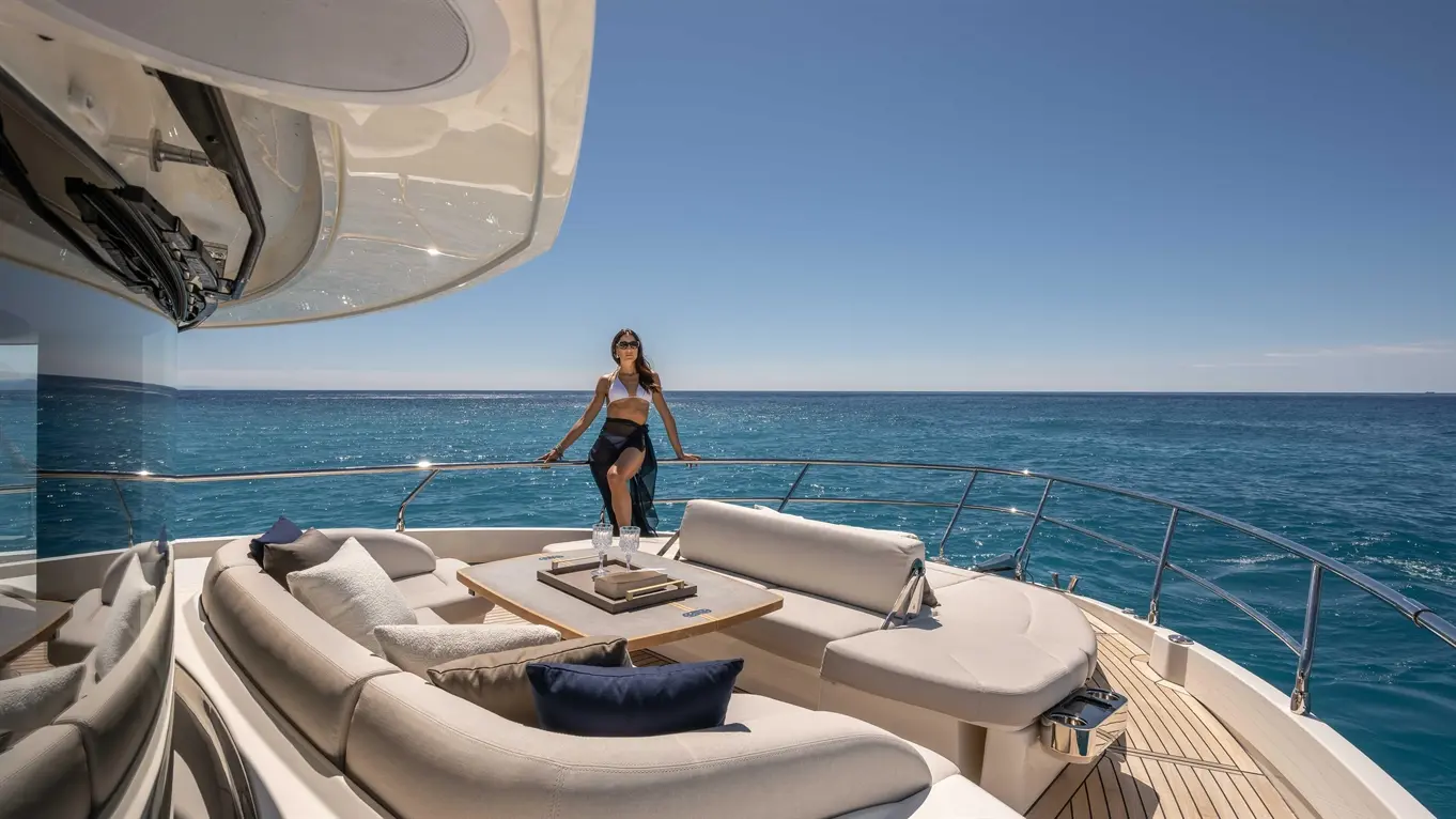 A woman in swimwear rests on a yacht's railing, overlooking calm blue waters, with plush seating and a table nearby.