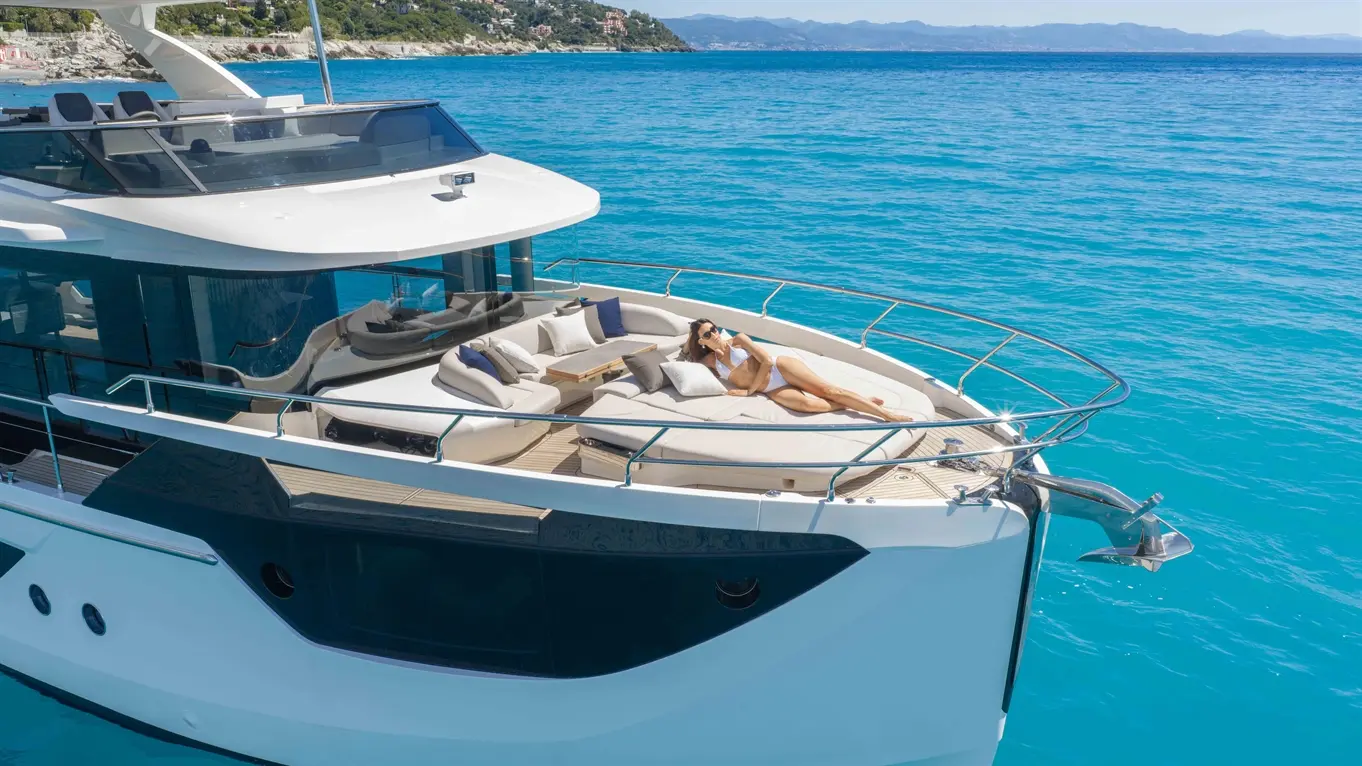A woman relaxes on a luxury yacht's sun deck, surrounded by cushions, against a backdrop of vibrant blue sea and coastline.