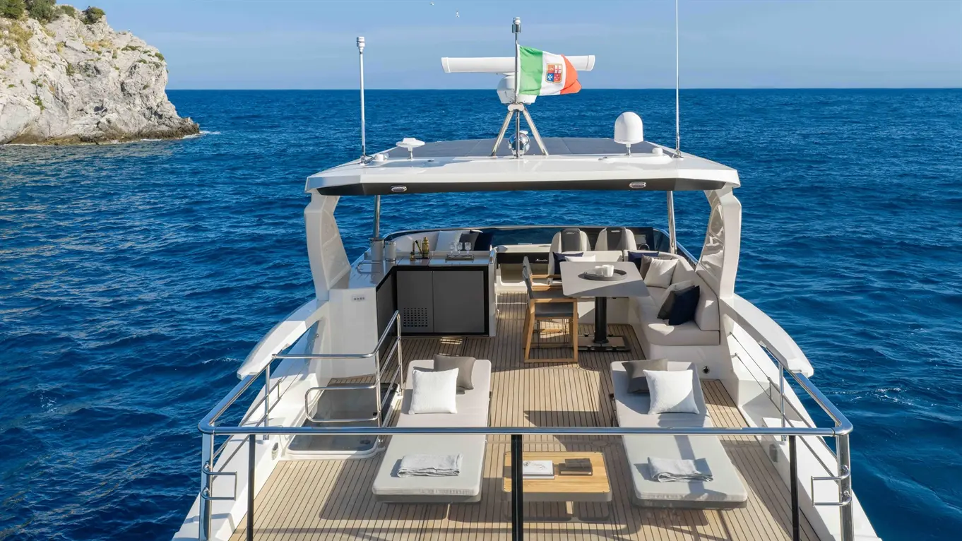 Luxury yacht deck with sun loungers, dining area, and Italian flag, set against a vibrant blue sea and rocky coastline.