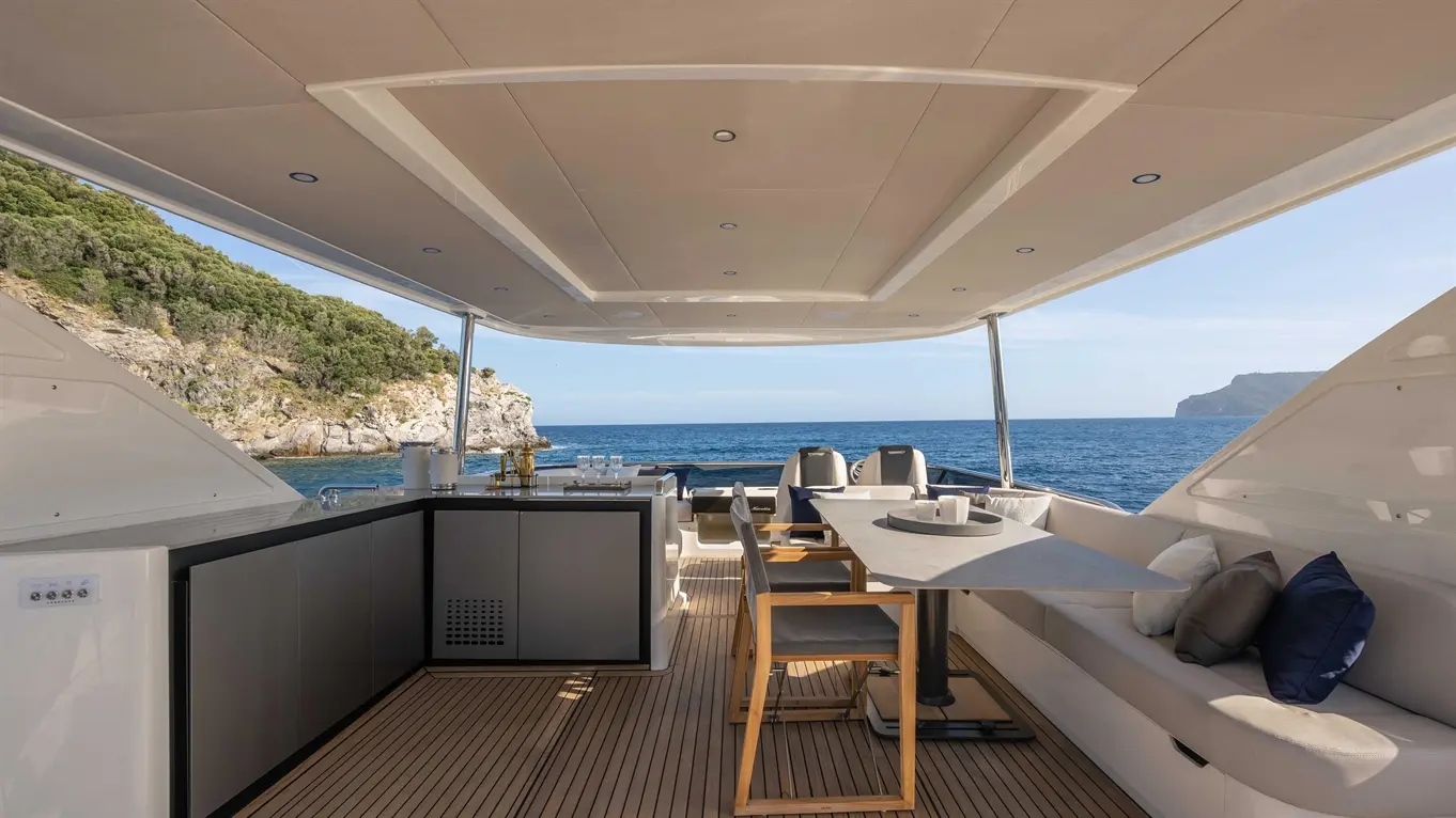 Luxury yacht interior featuring dining area, plush seating, and panoramic ocean views, with a rocky coastline in the background.