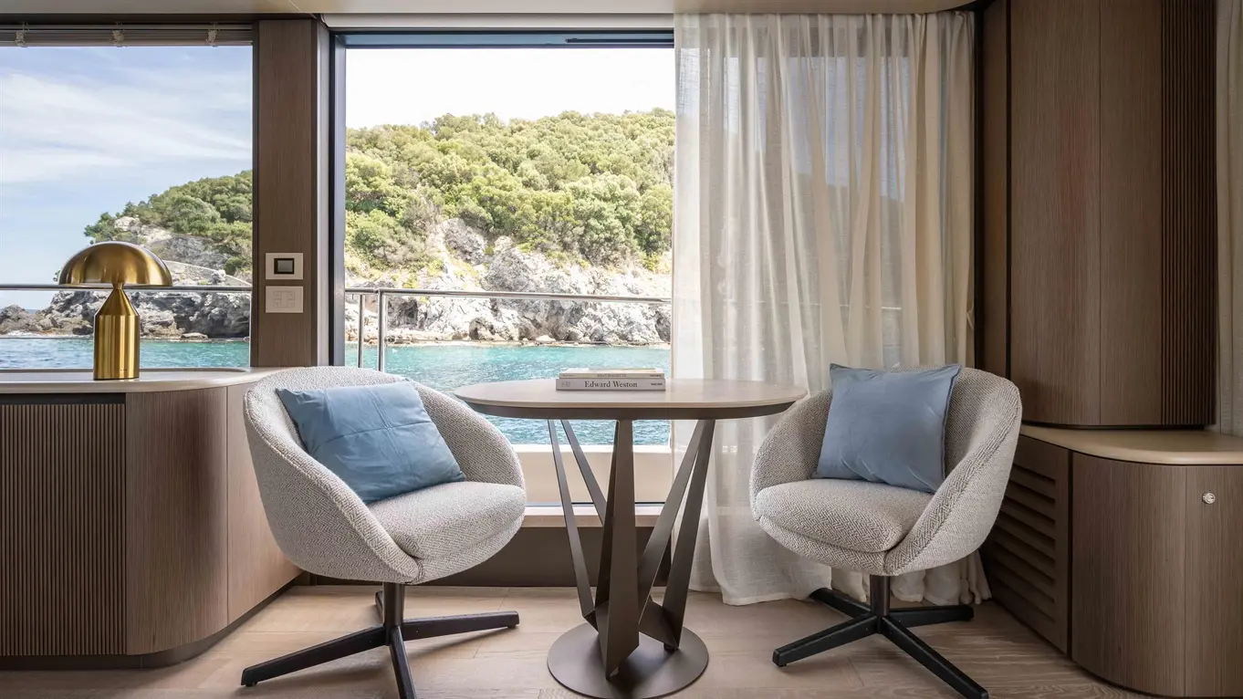 Modern lounge area with two plush chairs, a round table, a gold lamp, and a view of rocky greenery by the water.