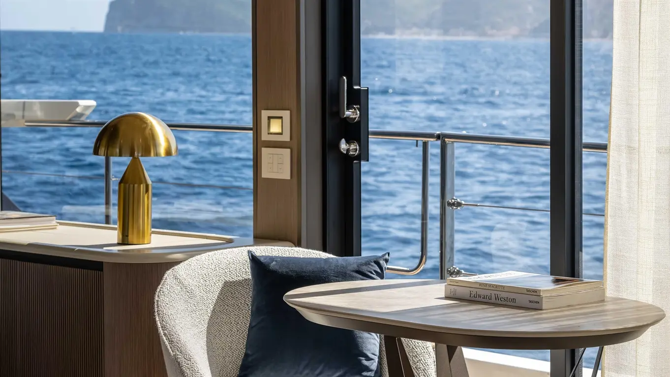 Modern yacht interior featuring a round table, stylish chairs, a golden lamp, and a view of the sea and mountains through glass doors.