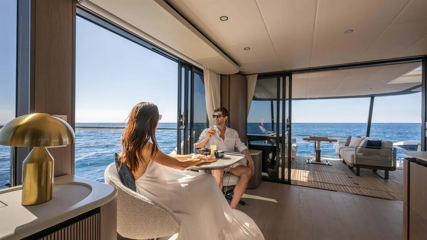 A couple enjoys brunch in a luxury yacht interior, with ocean views, sunlight, and modern furnishings.