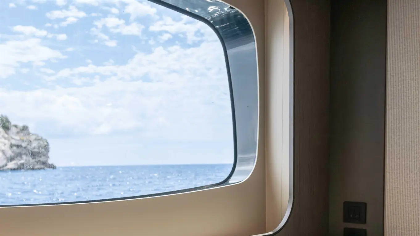 Interior view of a yacht showing two white cups on a surface with a large window overlooking the sea and cloudy sky.