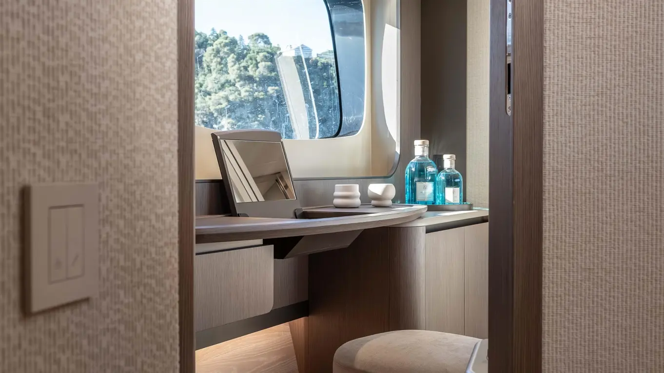 Luxurious yacht interior featuring a vanity, mirror, blue glass bottles, and a plush stool, with natural light from a large window.