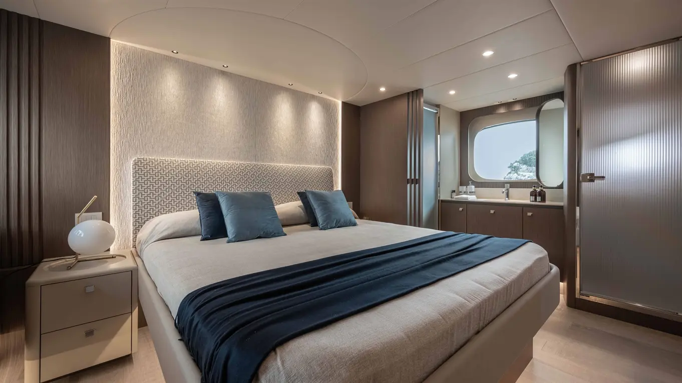 Modern bedroom featuring a large bed with blue pillows, bedside lamp, and a bathroom with a window and stylish decor.