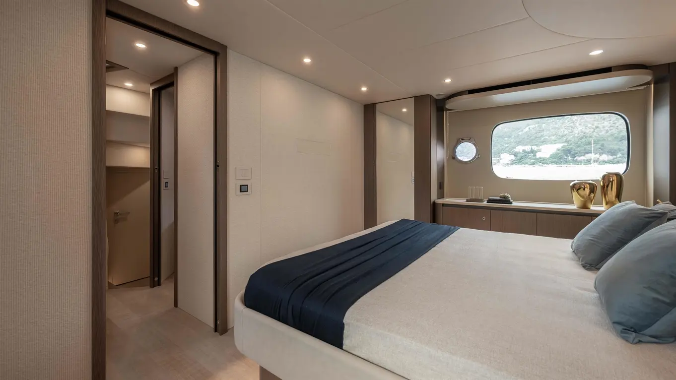 Modern yacht bedroom featuring a bed with navy bedspread, decorative pillows, and a round window view. Two stylish vases on shelf.
