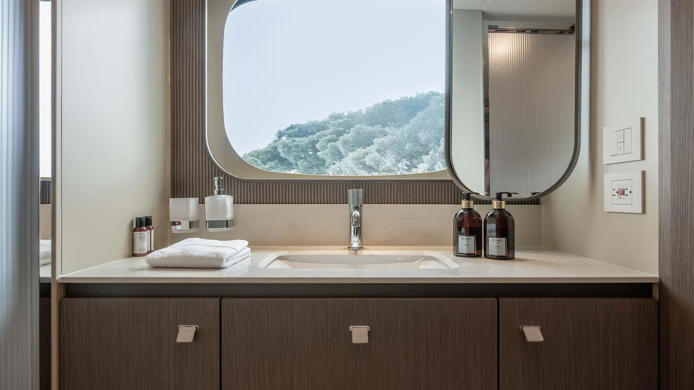 Modern bathroom with a sink, a large mirror, wooden cabinetry, towels, and toiletries, featuring a view of greenery outside.