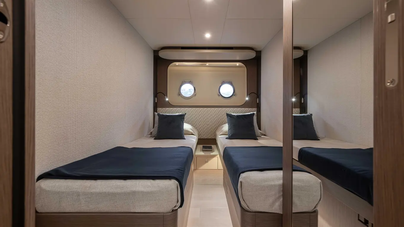 Stylish cabin with two single beds, blue throws, and pillows, illuminated by wall lights and featuring porthole windows.
