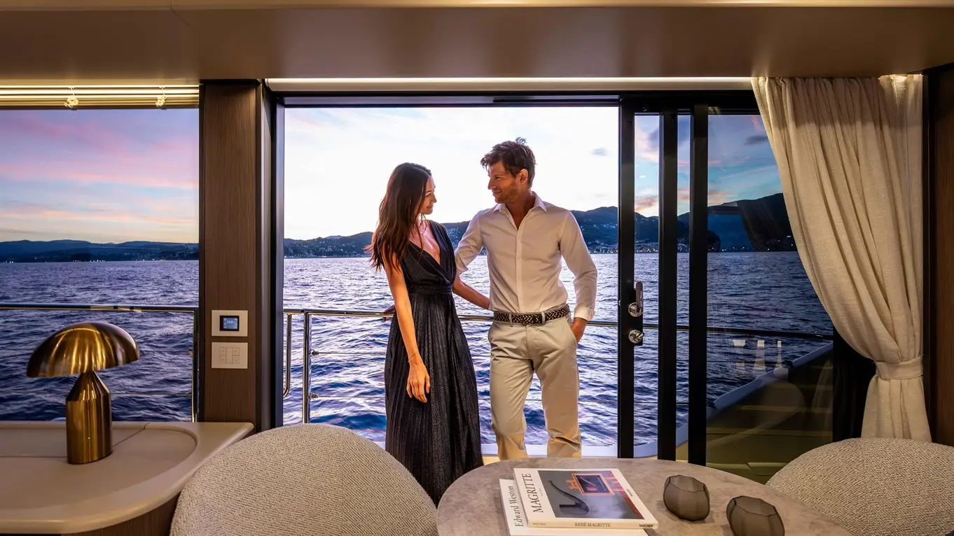 A couple stands by a large glass door on a yacht, with a sunset view over the water, surrounded by stylish interiors.
