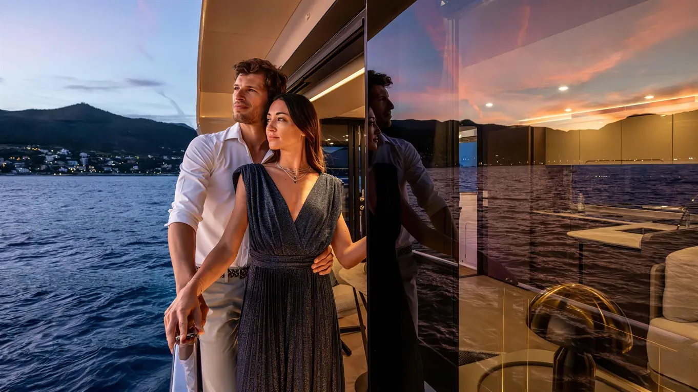 A couple on a luxury yacht during sunset, embracing with a view of mountains and water reflecting evening hues.