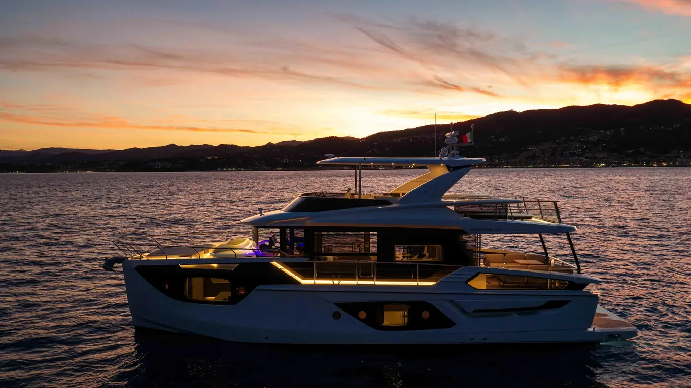 Luxury yacht anchored in water at sunset, featuring illuminated decks and a backdrop of silhouettes mountains and coastal lights.
