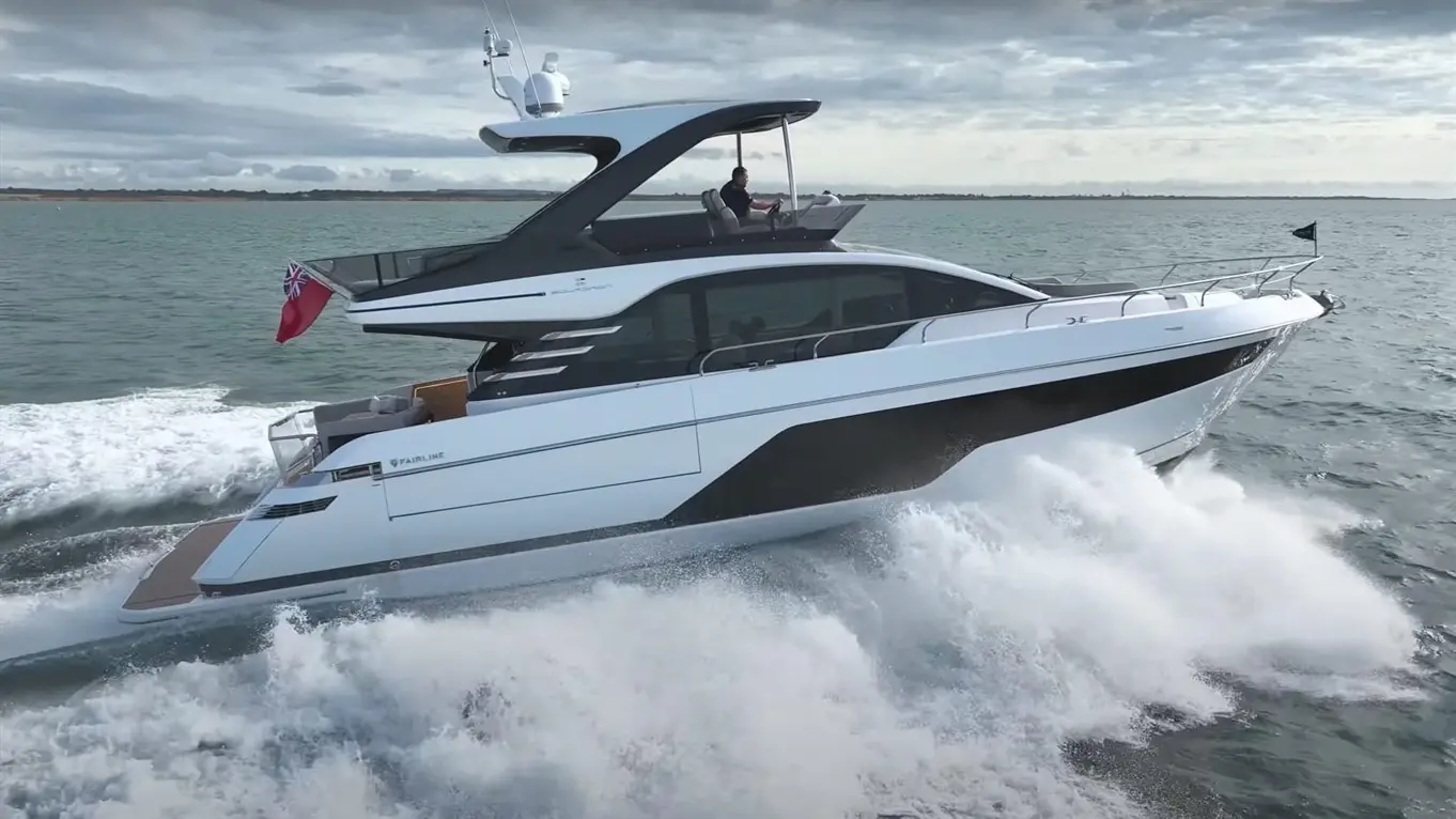 A sleek white yacht navigating through waves, with a Union Jack flag, and a man at the helm steering on a cloudy day.