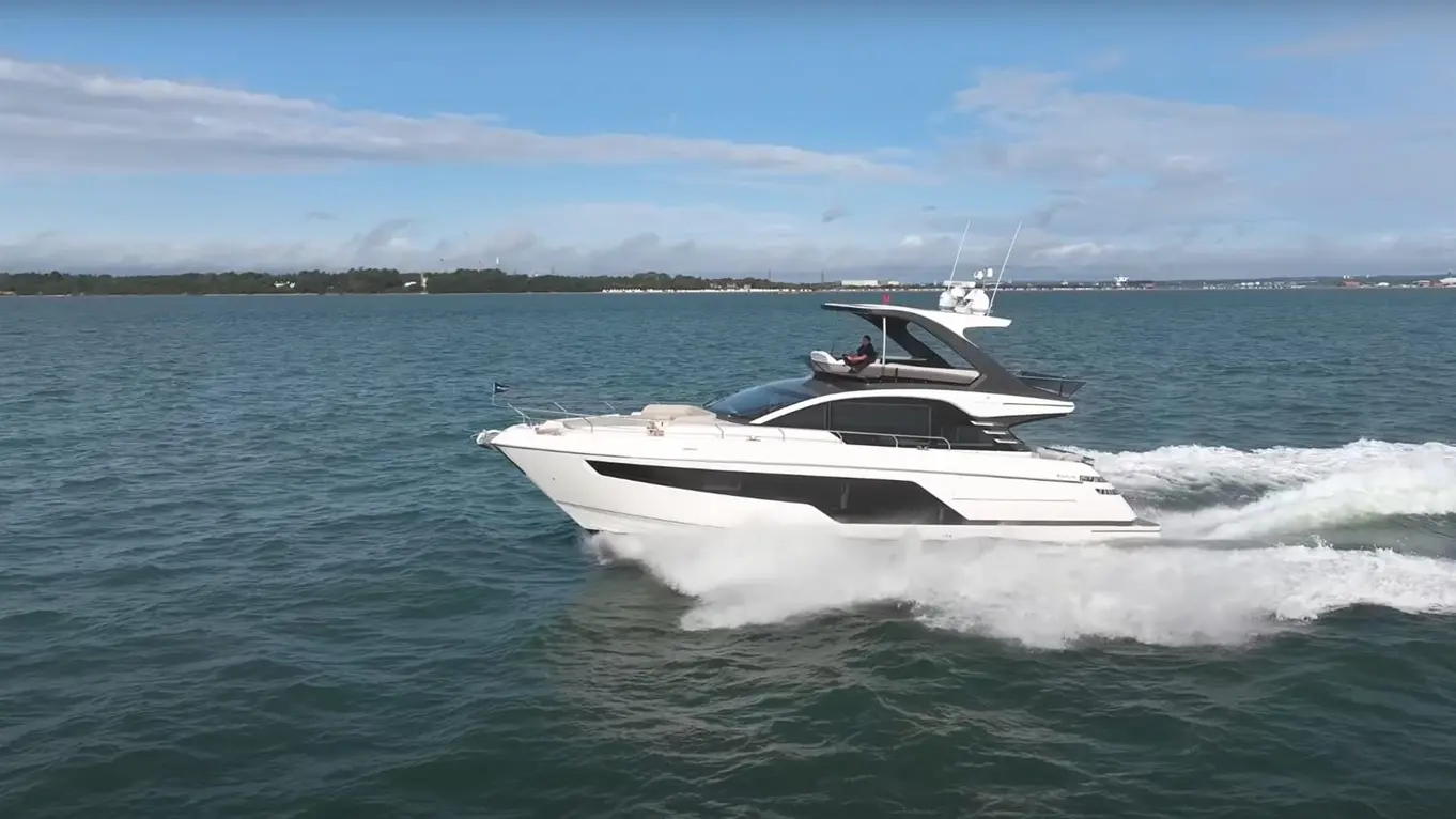 A sleek white yacht speeds through blue water, creating white foam waves under a partly cloudy sky.