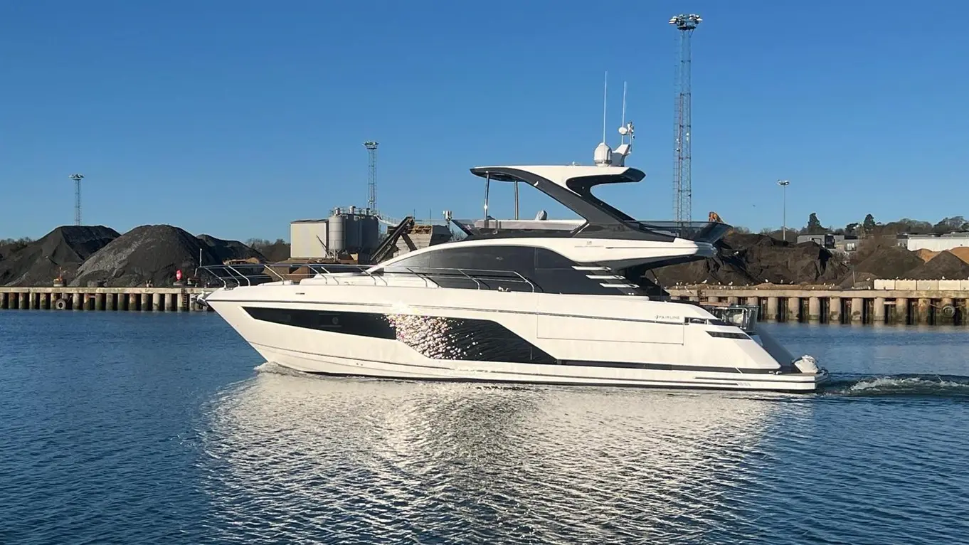 Luxury motor yacht cruising in calm waters, with a backdrop of industrial buildings and piles of material on the shoreline.