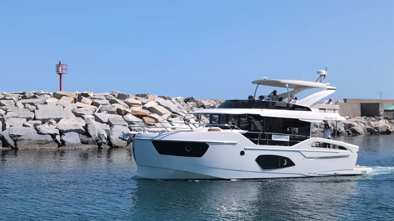 A sleek white yacht navigates calm waters near a rocky breakwater and a red navigational light under a clear blue sky.