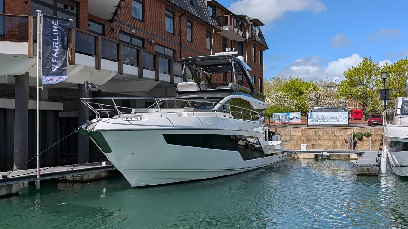 A sleek white Fairline yacht docked at a marina, surrounded by water and buildings under a blue sky with clouds.
