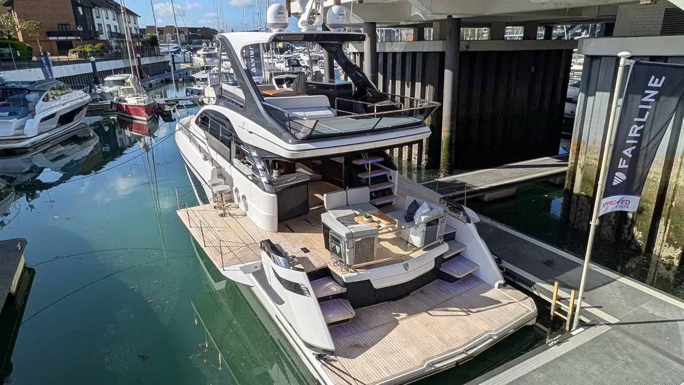 Luxury yacht in marina with plush seating, wooden deck, and Fairline flag visible, surrounded by moored boats and clear water.