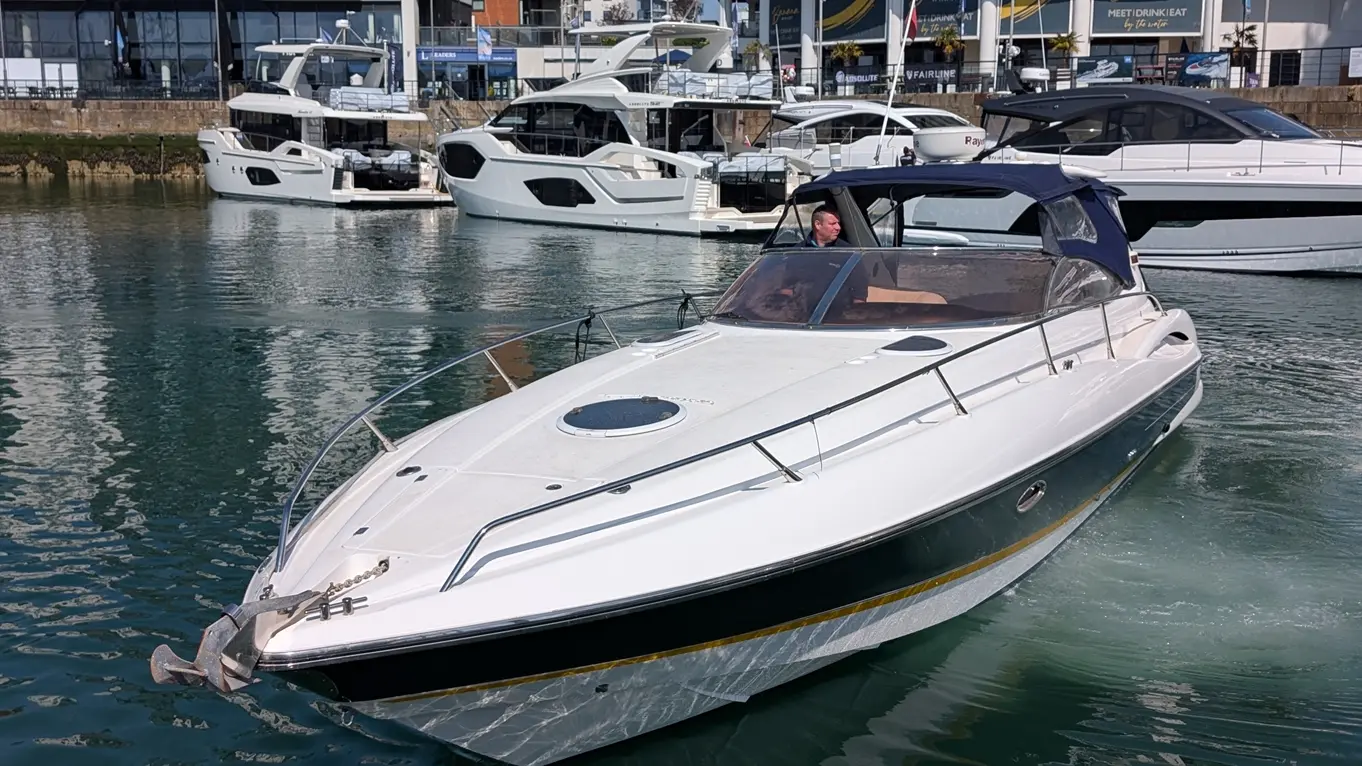 A white motorboat navigates calm waters near luxury yachts, with a man steering at the helm and marina buildings in the background.