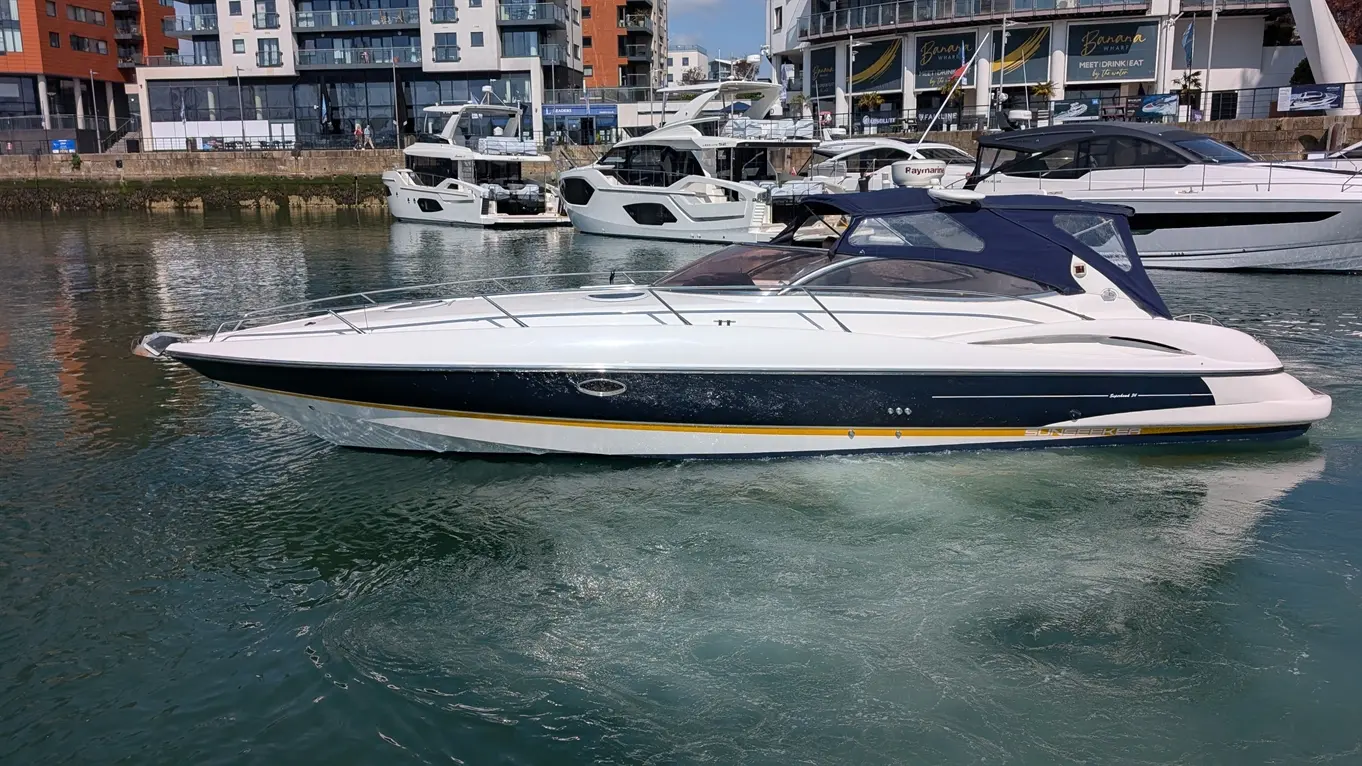 A sleek white and navy motorboat with a blue canopy sails through clear water, surrounded by luxury yachts and modern buildings.