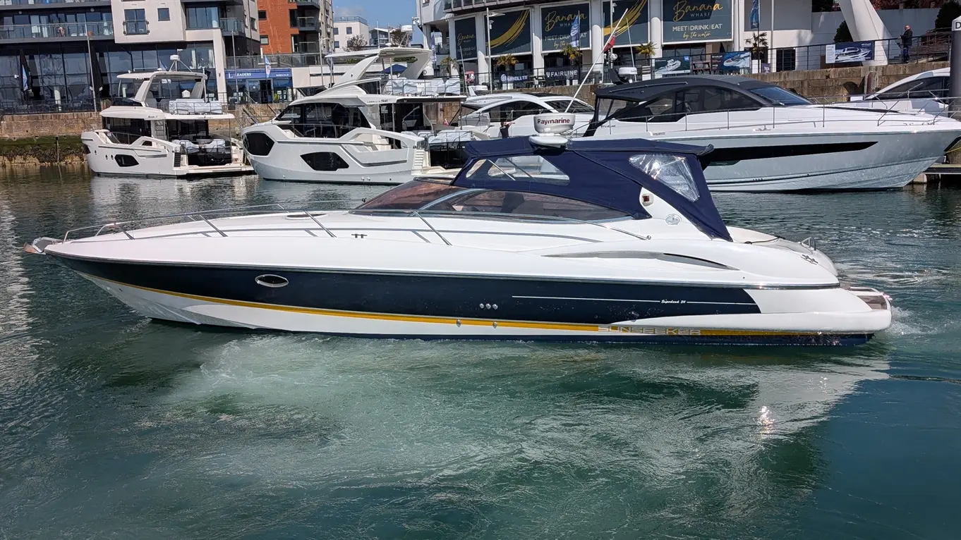A sleek motorboat navigates a marina with luxury yachts, featuring a white and navy design and a "Sunseeker" logo.