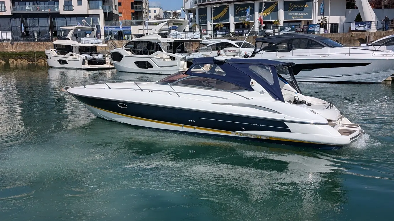 A sleek white and blue Sunseeker boat is moored in clear water, surrounded by luxury yachts and buildings at a marina.