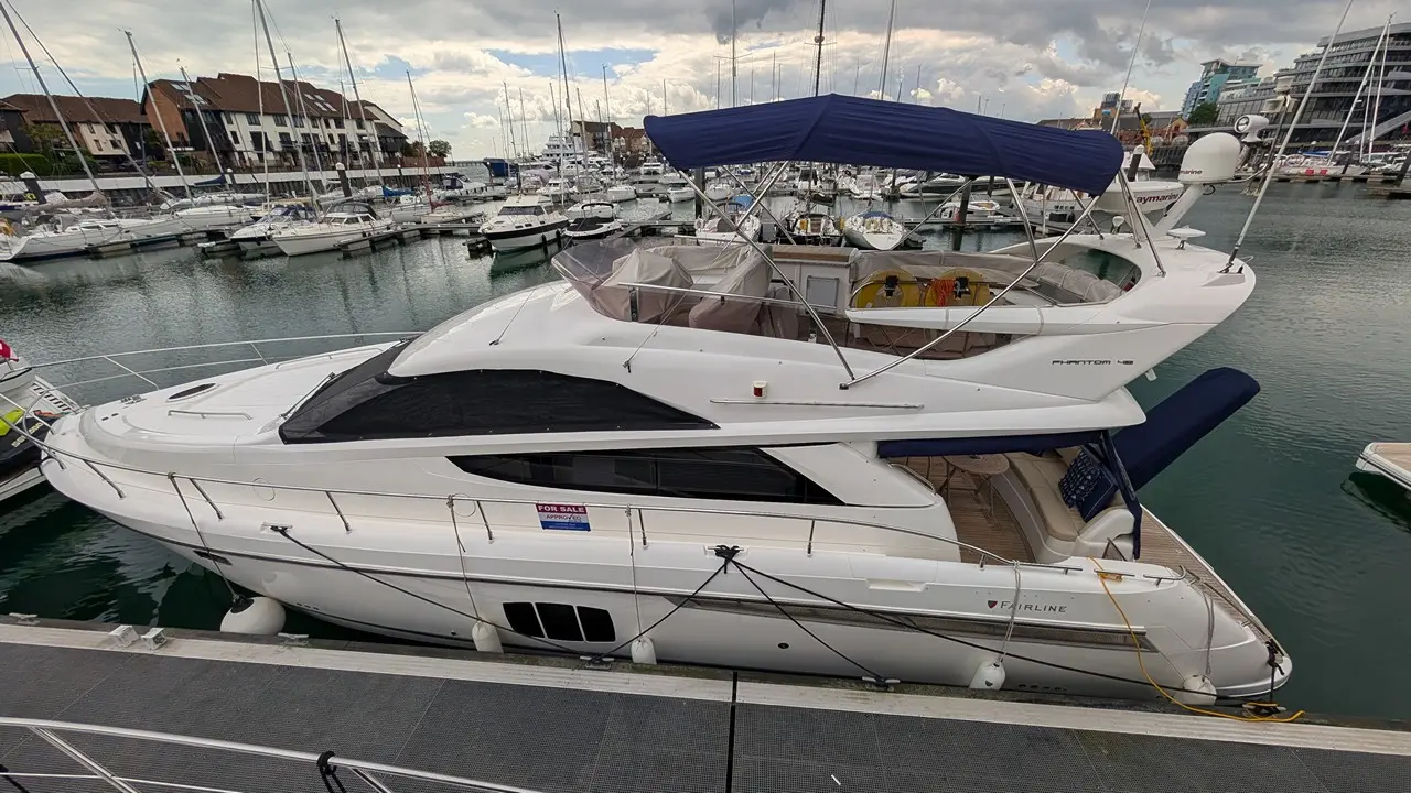 A white luxury yacht moored in a marina, featuring a 'FOR SALE' sign, surrounded by other boats and a cloudy sky.