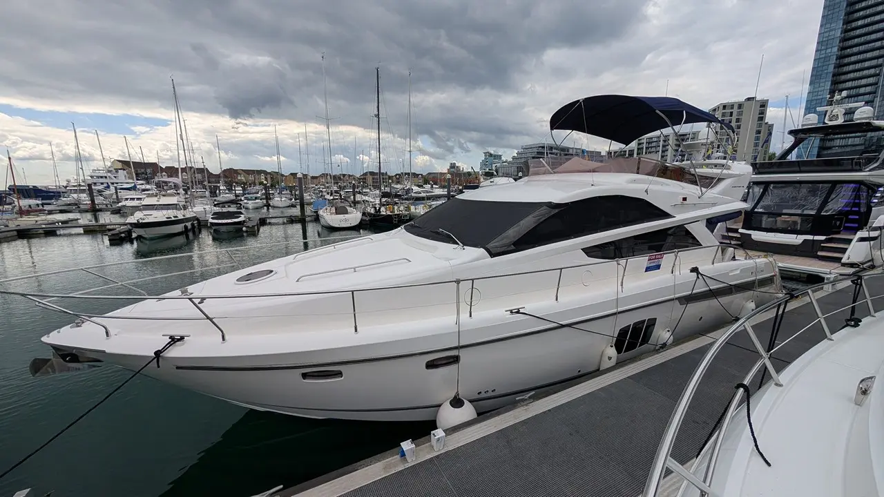 A luxury motor yacht docked at a marina, with a "For Sale" sign and a backdrop of cloudy skies and other boats.