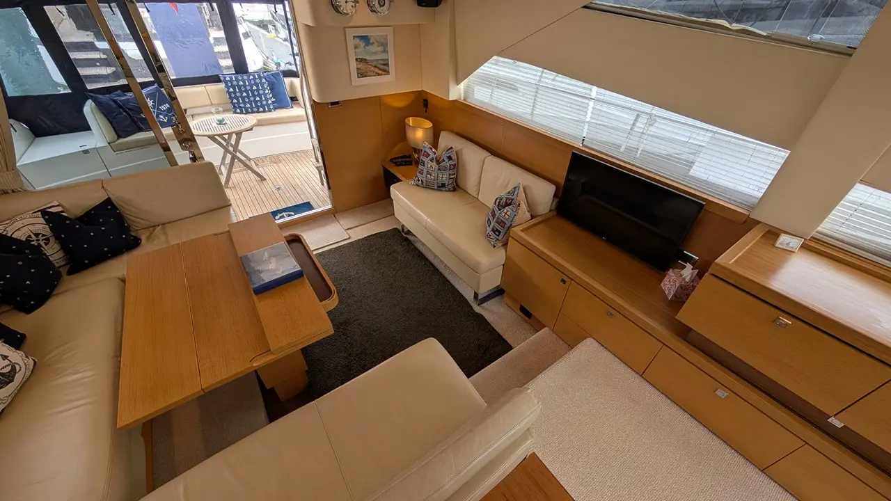 A stylish yacht interior featuring a sunlit lounge, cream sofas, decorative cushions, a wooden table, and a TV unit.