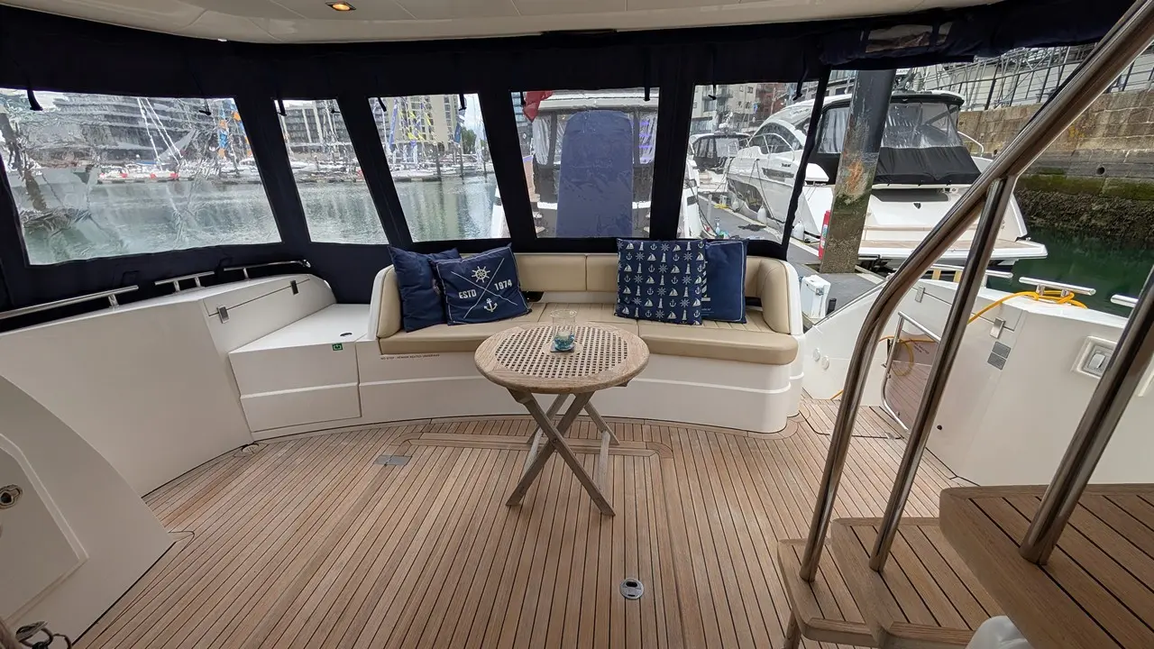 Interior of a boat featuring a comfortable L-shaped seating area, wooden floor, and a round table, overlooking a marina.