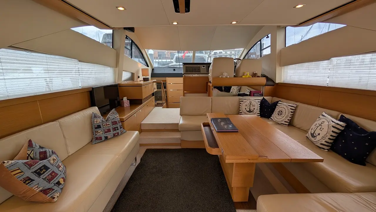Stylish yacht interior featuring cream leather seating, wooden furniture, a dining table, and decorative pillows by large windows.