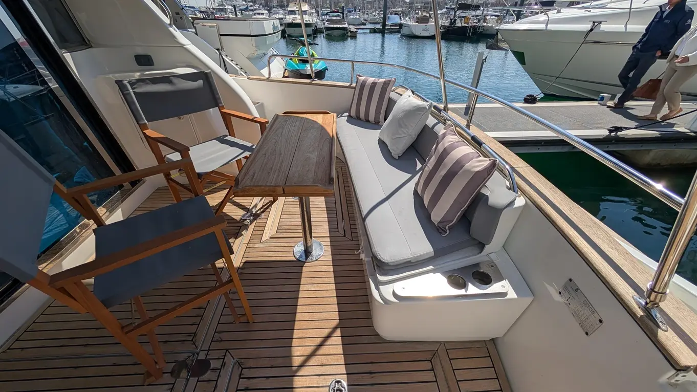 A sunny yacht deck featuring grey cushioned seating, wooden table, and marina views with moored boats in the background.
