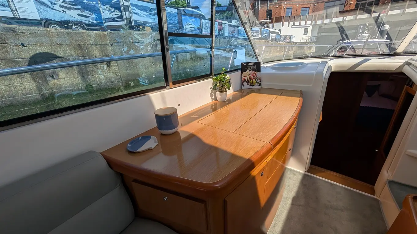 A bright interior of a boat features a wooden table, a plant, and a cup, with a view of boats through large windows.