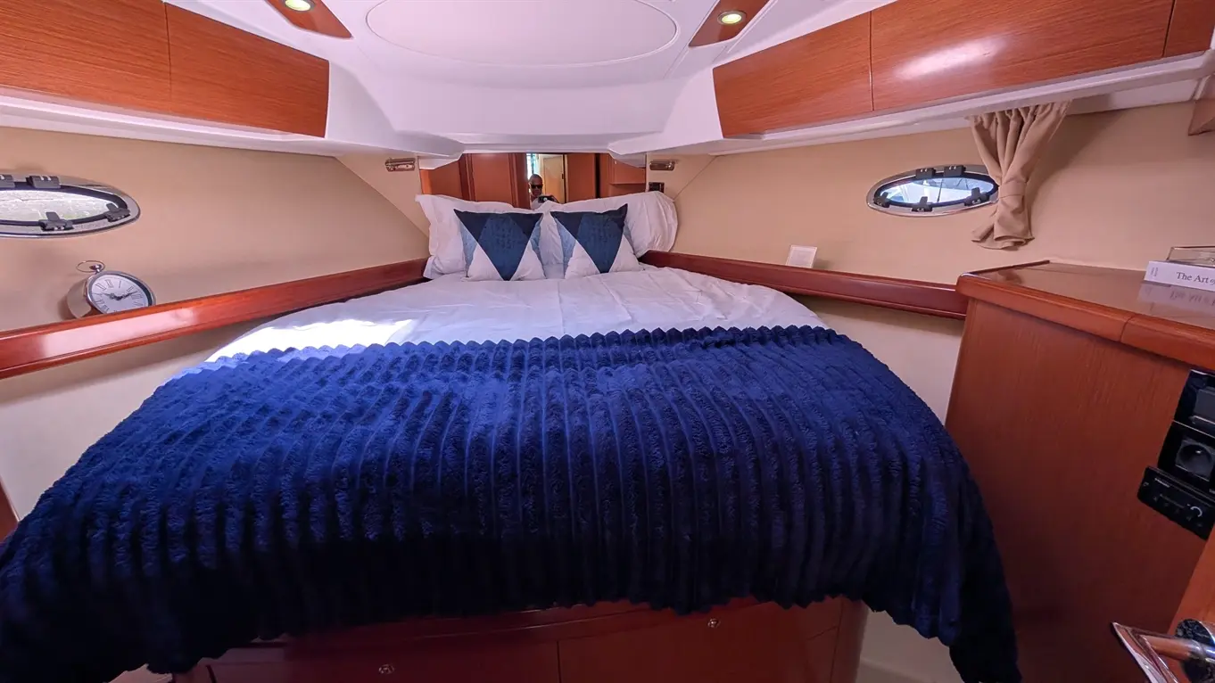 Cosy cabin featuring a bed with a navy throw and geometric pillows, wooden cabinets, and an oval window with a clock.