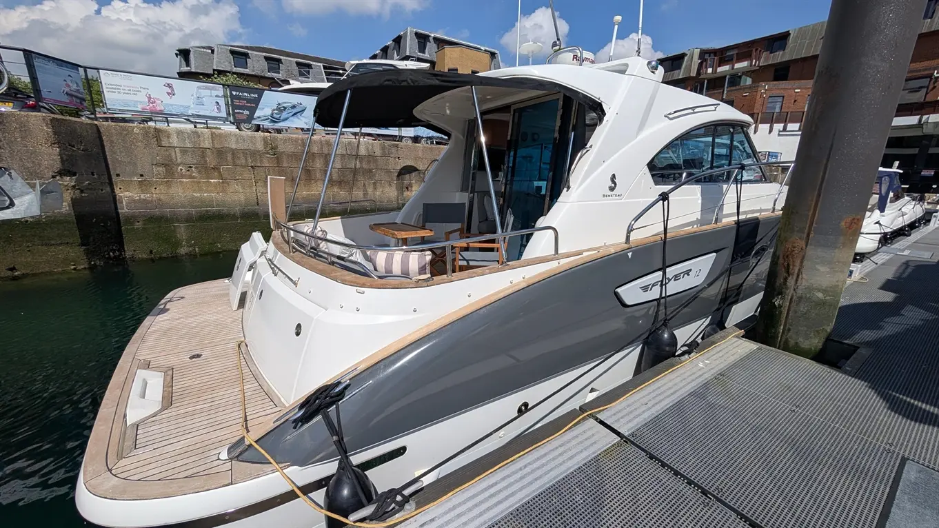 Luxury motorboat docked by a harbour, featuring a wooden deck, seating area, and modern design under a blue sky.