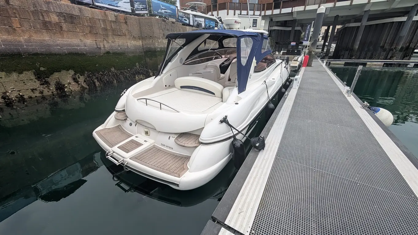 A white motorboat moored alongside a jetty, with wooden decking and a blue canopy, reflecting in calm water.