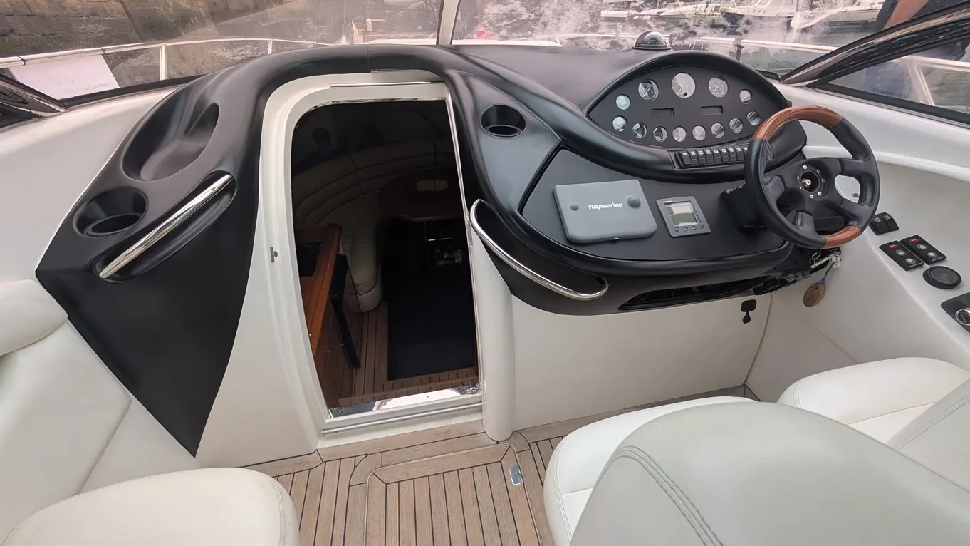 Interior view of a boat with a steering wheel, dashboard controls, and an entryway to a cabin featuring wood accents.