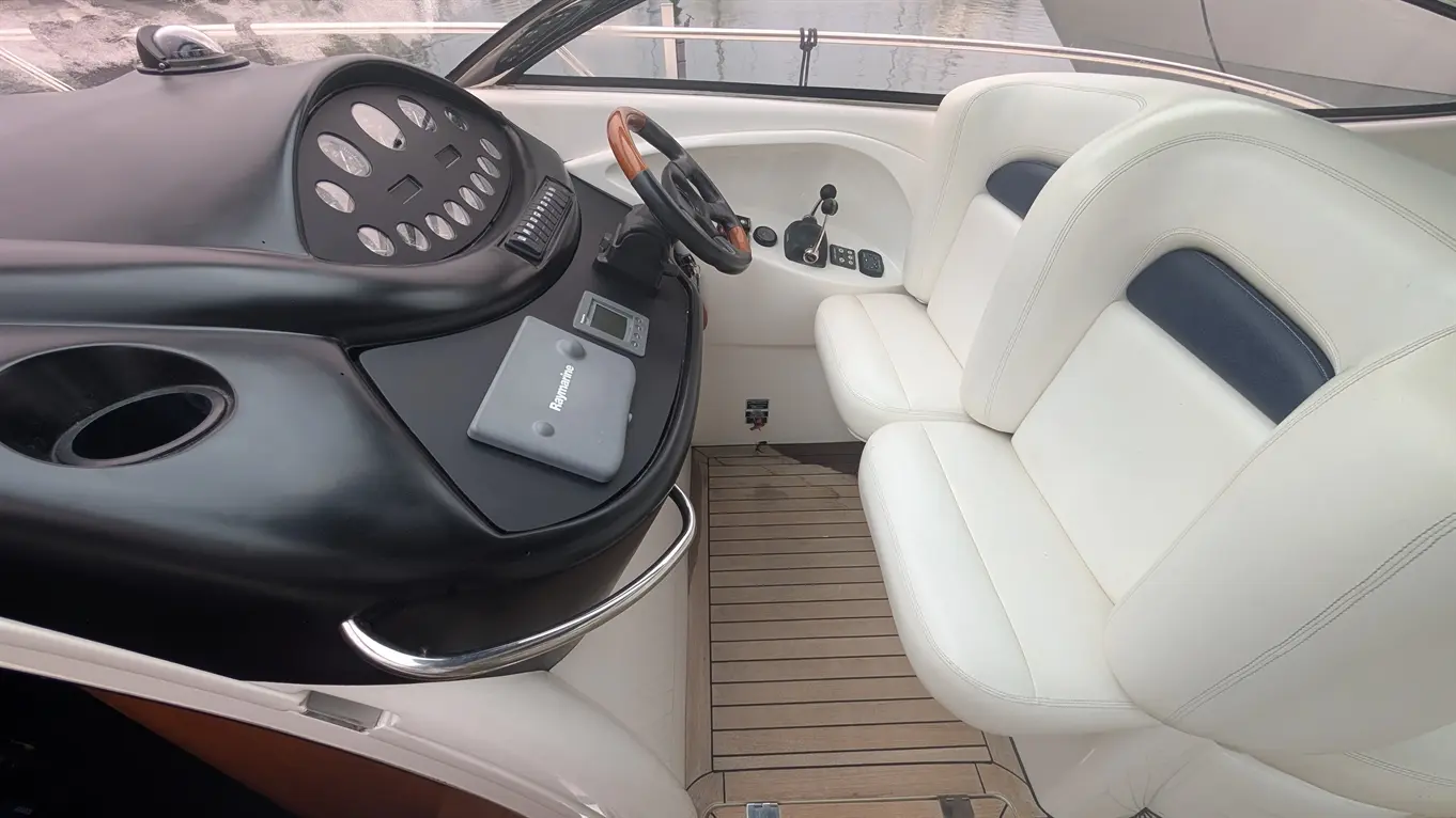 Boat cockpit featuring a steering wheel, instrument panel, and white leather seats with wooden flooring. Raymarine device visible.