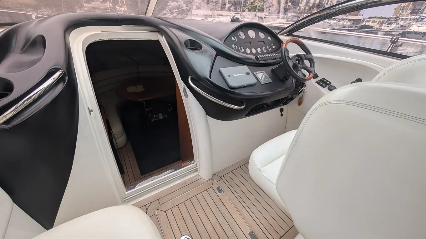 Interior view of a boat, featuring a sleek dashboard, steering wheel, and white seating, with a cabin entrance visible.