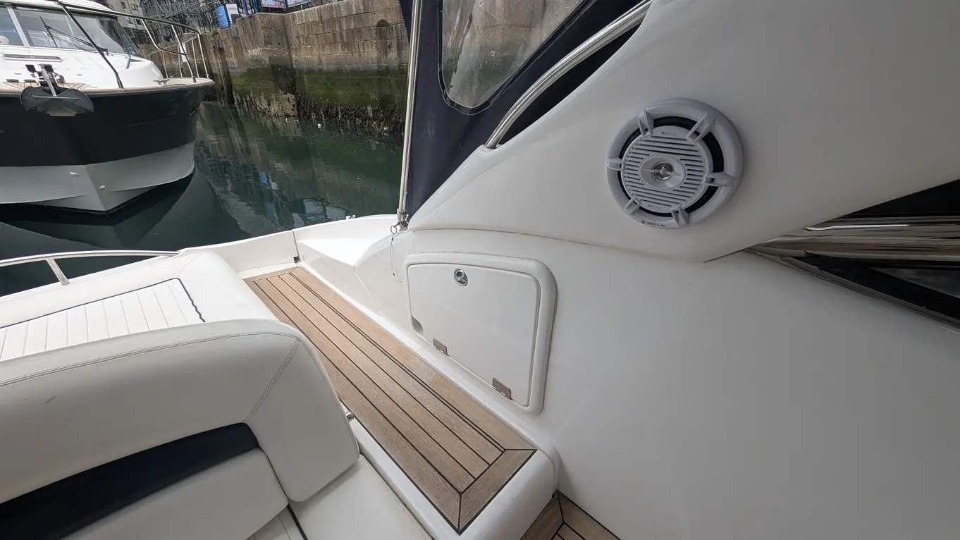 Interior of a boat showing white seating, wooden decking, a speaker, and the reflection of a nearby docked vessel.
