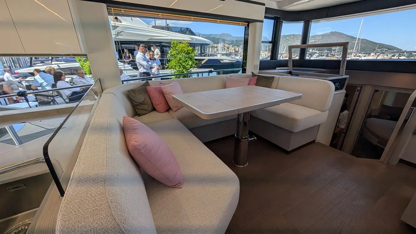 Luxury yacht interior with comfortable seating, pink cushions, and a table, overlooking marina and mountains through large windows.