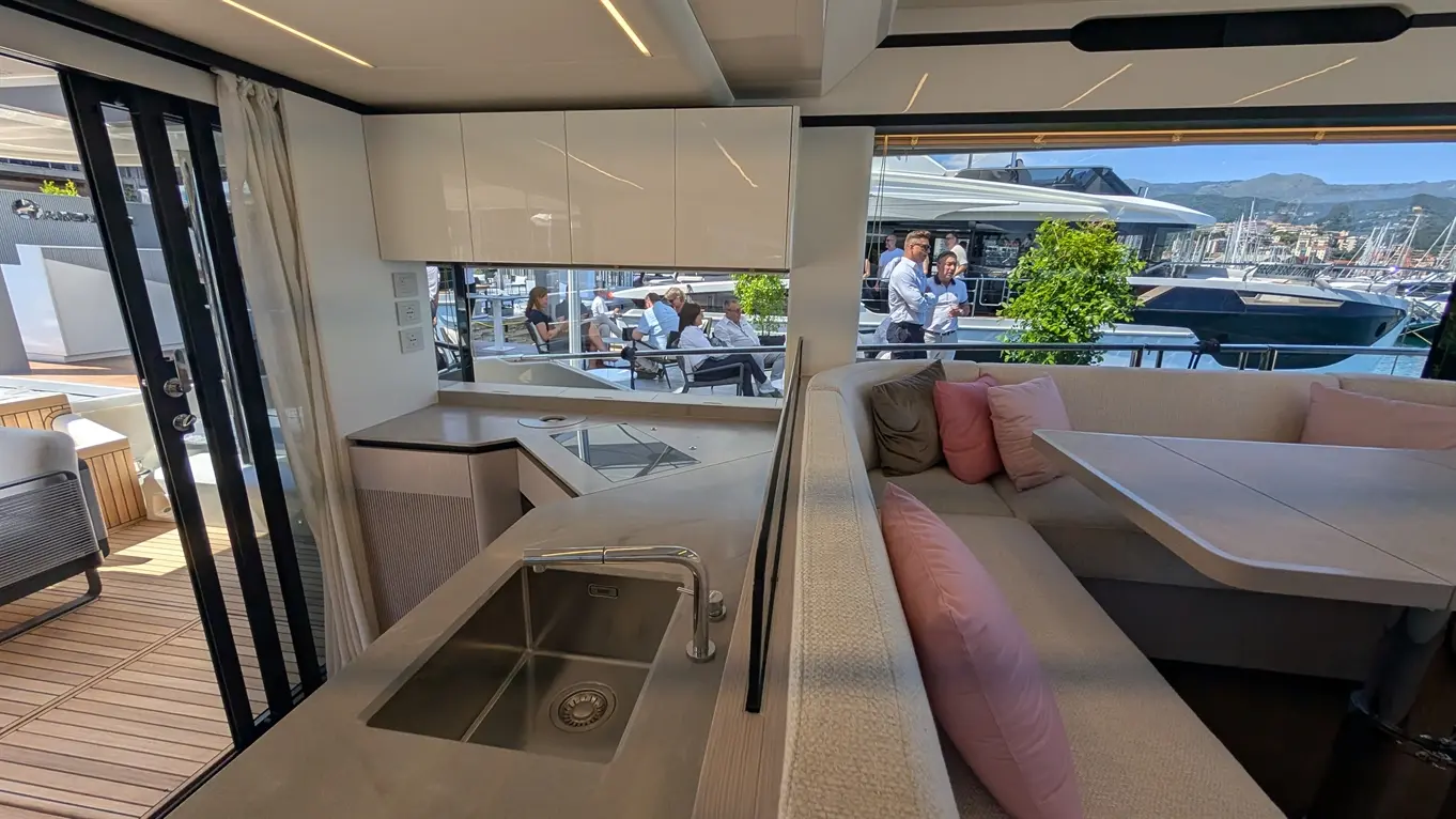 Interior of a luxury yacht with a modern kitchen, plush seating, and a view of people socialising by the marina.