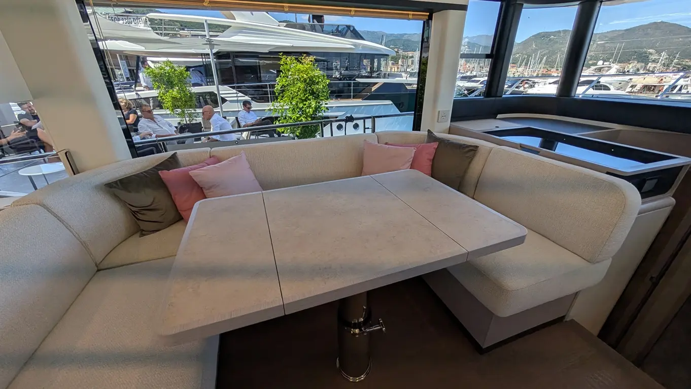 Stylish yacht interior with a large dining table, plush seating, and views of boats and greenery through the windows.
