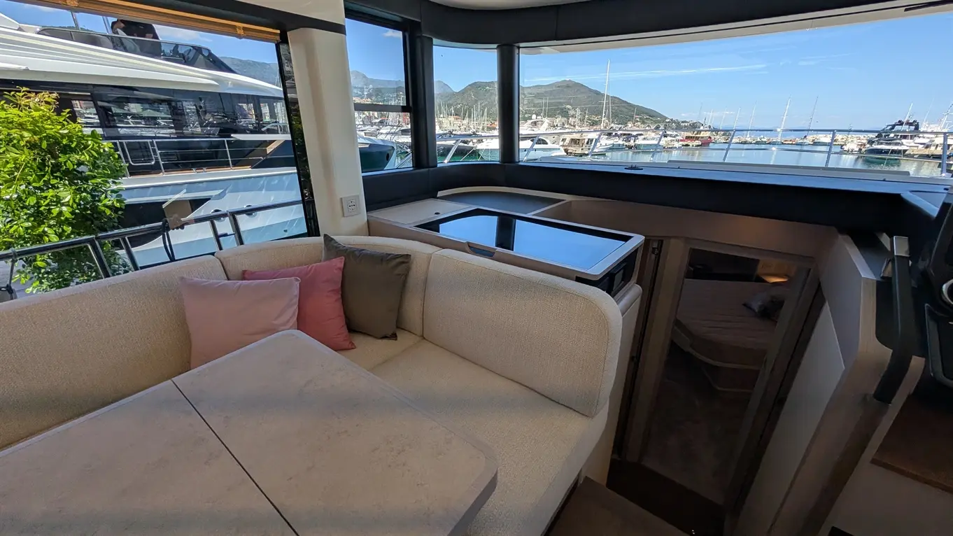 Interior of a yacht featuring a plush seating area, table, and large windows with views of a marina and mountains.