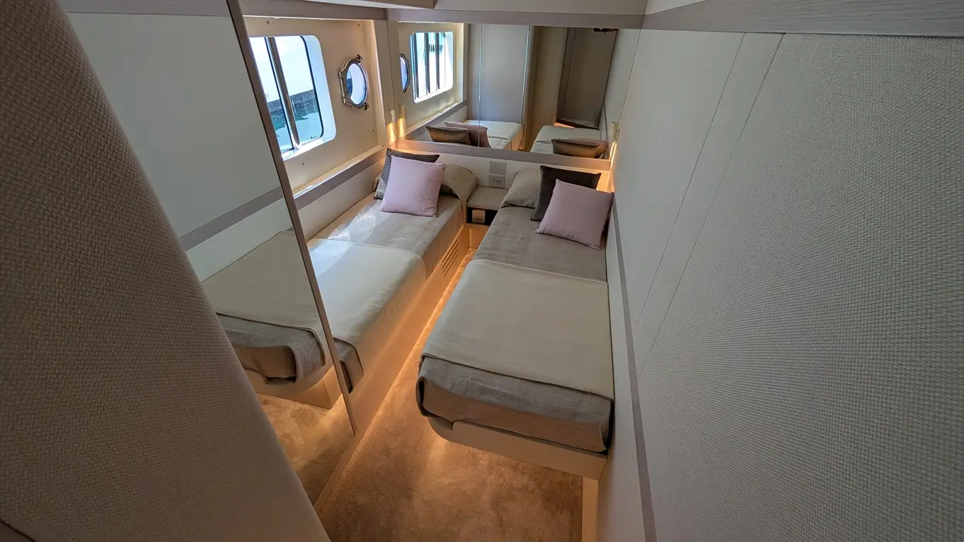 Modern boat cabin with two single beds dressed in light linens and pillows, featuring mirrors and round windows for natural light.